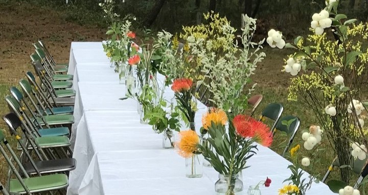 野原に置かれた長いテーブルの上に、真っ白なテーブルクロスがかけられ、たくさんの花瓶に花が飾られており、周囲に椅子がたくさんおかれている風景写真